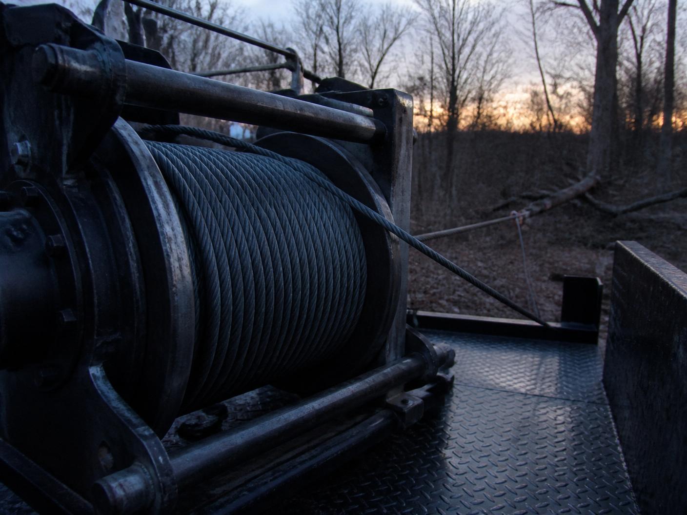 Heavy steel hydraulic winch drum with wire cable spooled on a work truck bed, cable running to a fallen tree at dusk